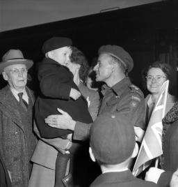 A black-and-white image of a smiling man in a Canadian army uniform smiling and talking to a small child who is being held by a woman. Other men and women are gathered around.