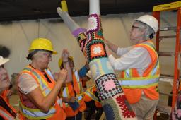 Women wearing hard hats and orange, white and yellow safety vests place yarn squares on a replica of a tree branch.