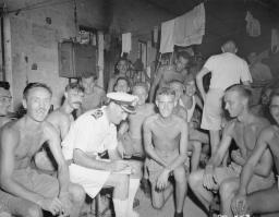 A black-and-white image of more than a dozen men gathered in a small, dark room. Many are smiling at the camera. Most are shirtless and are very emaciated.