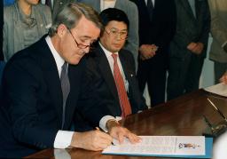 Brian Mulroney sitting and signing a document. Art Miki is sitting beside him looking on.