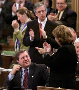 In the House of Commons, a man is sitting while his fellow politicians are standing and applauding.