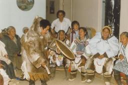 A man dancing and playing an Inuit drum while a group of elderly women sings.