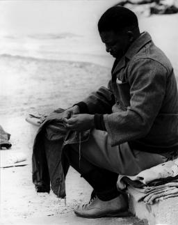 A black-and-white image of Nelson Mandela. He is sitting and holding a piece of clothing on his knees. He appears to be sewing. He is wearing shorts, long socks, white shoes and a jacket.