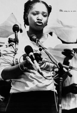 A black-and-white photo of Zindzi Mandela, a young black woman, standing and speaking in front of several microphones.