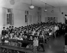 Row upon row of children sit on a long row benches in a large room. 