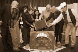 A group of people standing around the Truth and Reconciliation Commission's bentwood box. They are leaning towards the box and reaching with their hands to lower a suitcase in the box.