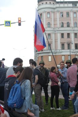 Two soldiers in uniform stand behind a crowd of young people. Two of the young people are holding a red, white and blue striped flag. There is a tall building in the background.