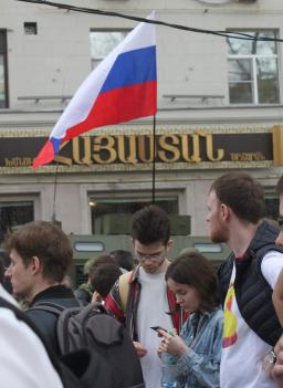 Amid a crowd of milling people, a young woman and a young man look down at the mobile devices in their hands. Directly behind them is a red, white and blue striped flag.