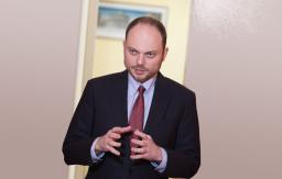 A man with a short beard and moustache, wearing a suit, talks with his hands held in front of him. 