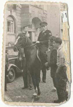 Black and white photo with tattered edges showing a uniformed man on horseback in the street. Two men are standing nearby. An old car is in the background.