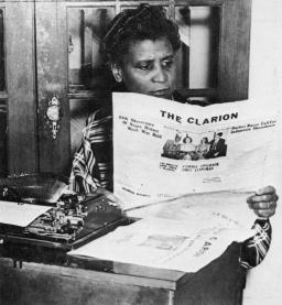 A black and white image of a Black woman sitting in front of an old-fashioned typewriter, reading a newspaper named “The Clarion”.