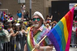 A man with sunglasses and a rainbow-coloured shirt carries a large Pride flag while a crowd of onlookers watch from a distance. 