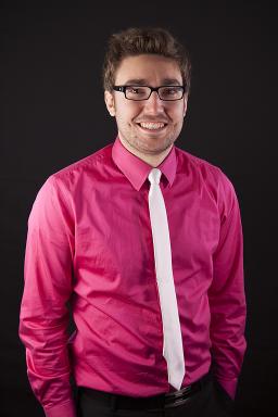 A man wearing a pink shirt and a white tie looks at the camera and smiles.