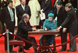 Former Prime Minister Pierre Elliott Trudeau sits to the left of Queen Elizabeth II at a wooden table. Two men lean over her right shoulder as she signs a document.