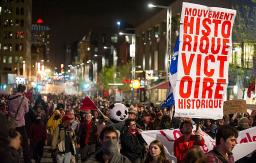 A sea of young adults marching down a busy city street in the evening, with buildings and lights surrounding them. One person is holding a sign that says “historic movement, historic victory” in French. Another is wearing a large panda bear mask.