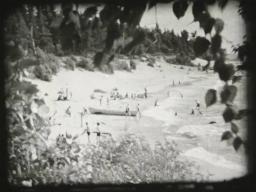 A black-and-white photograph of a beach scene, with people on the sand and in the water, and a canoe on the sand near the water.