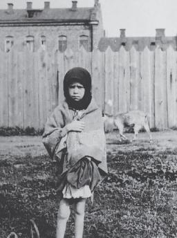 A young girl stands facing the camera with a sorrowful expression, clutching a shawl around her.