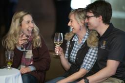 Three laughing people sit together at a table in a Museum space, drinking wine.