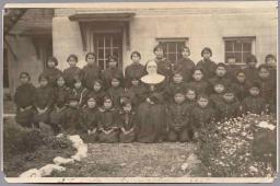 A nun and a group of First Nations children in school uniforms posing in front of a stone building.