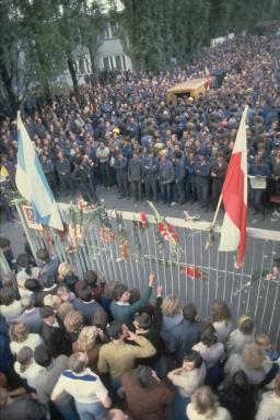 Thousands of people wearing blue uniforms are crowded on one side of a high gate. People in everyday clothing appear on the other side of the gate, which is decorated with flowers and two flags.