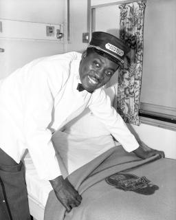 A uniformed man making a bed in a railway sleeping car berth.