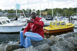 A man wearing a brightly coloured track suit and a white skullcap sits on some rocks, with boats and a harbour in the background.