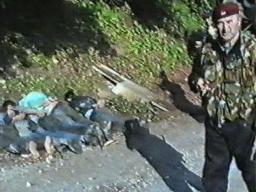 Grainy still image from a video showing a man wearing a camouflage uniform in the foreground. Several people in civilian clothes lie face down in the background with their hands tied. 