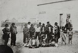 Black and white photograph of a group of men standing and sitting in front of a building with the word “Saloon” painted prominently on the wall.