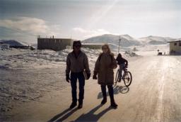 Two people in winter coats stand in a snowy landscape with hills and buildings in the distance.