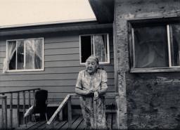 A white-haired woman leans on a porch railing outside a house.