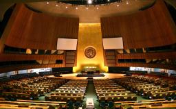 A large hall with rows of desks and chairs facing a high wall emblazoned with the UN logo.