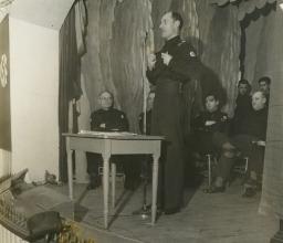 A man wearing a uniform bearing a swastika speaks into a microphone while a group of similarly uniformed men look on.