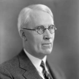 Formal portrait of a white-haired man wearing glasses and a three-piece suit. 