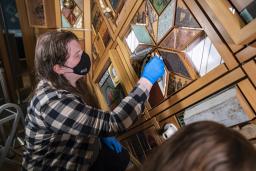 A person with long hair, wearing a face mask and blue gloves, cleans a section of a wood-framed artwork with a small sponge.