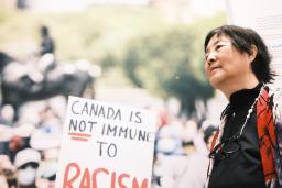 A woman stands next to a large photo of a crowd where someone is holding a sign that reads “Canada is not immune to racism.”