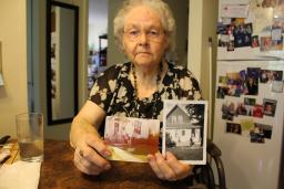 An elderly woman sits at a kitchen table and holds up a colour photograph of a family sitting on a front porch and a black-and-white photograph of a house with a woman sitting on the front steps. 