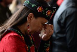 A person with long hair and beaded earrings and headgear bows their head and holds a tissue to their eyes.