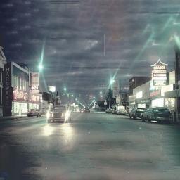 A blurry photograph of a 1970s-era small-town main street at dusk, with bright neon signs and streetlights.