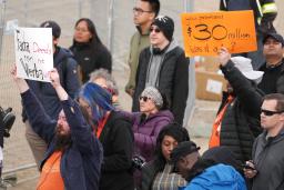 A crowd of people standing outside, some holding signs reading “Facta non Verba: Deeds not words” and “You promised $30 million. Was it a lie?”