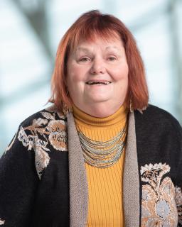 A white woman with red hair and brown eyes wears a yellow turtleneck and black cardigan with grey-brown flowers. She's front-facing and smiling at the camera.