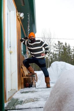 a Sikh-Canadian man, smiling, wearing a black and white striped sweater, blue and brown pants, boots, and an orange turban, in the snow is leaning on the side of a cabin.