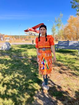 A young woman is shown wearing an orange jingle dress. She is outside in a park. An airplane statue can be seen behind her. 