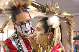 Two people dressed in traditional Indigenous regalia. One person is painting the other person’s face with black and white markings.