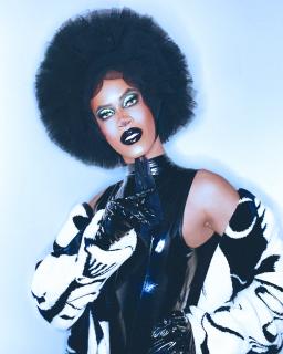 A biracial man dressed in drag. He is wearing a black headpiece, black leather gloves, a black leather dress and and black and white sweater.