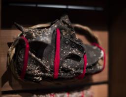 An old, broken child’s leather shoe covered in lichen. It has been loosely wrapped with a strip of red cloth, mounted on a wood backing and encircled with a braid of sweetgrass.