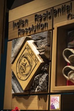 A square piece of wood printed with a drawing of a face mounted on a wooden surface and surrounded by pieces of raw, worn, and weathered wood. Next to it is a glass-fronted display case containing leather moccasins.