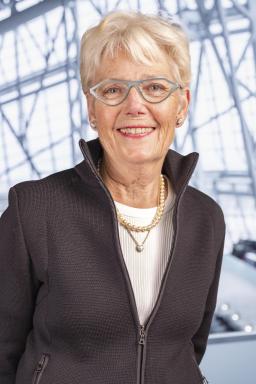 A white woman with short blond hair is standing with her arms behind her back in a relaxed manner. She is wearing a black sweater, a white shirt and blue-grey eyeglasses.