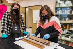 Two people wearing face masks and blue gloves stand at a worktable in front of a large glass cabinet containing many bottles and jars. One person is working on one of two oblong pieces of wood laid out on protective paper as the other person looks on.