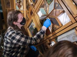 A person wearing a face mask and blue gloves using a small sponge to clean a section of a large sculptural artwork consisting of many elements made of wood and other substances arranged in interlocking geometric patterns.