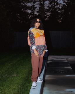 Person with curly shoulder-length dark hair is standing in a garden at dusk with sunglasses. They are wearing a colourful outfit.